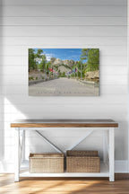 A rectangular wall art print showing a scenic mountain road with trees, flags, and a blue sky, displayed above a white console table with a wood top in a bright room with white shiplap walls.