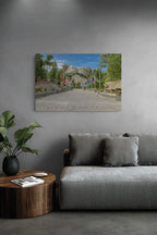A rectangular wall art print showing a scenic road leading up a mountain with trees, flags, and rocky peaks under a blue sky, displayed above a gray sofa in a modern living room setting.