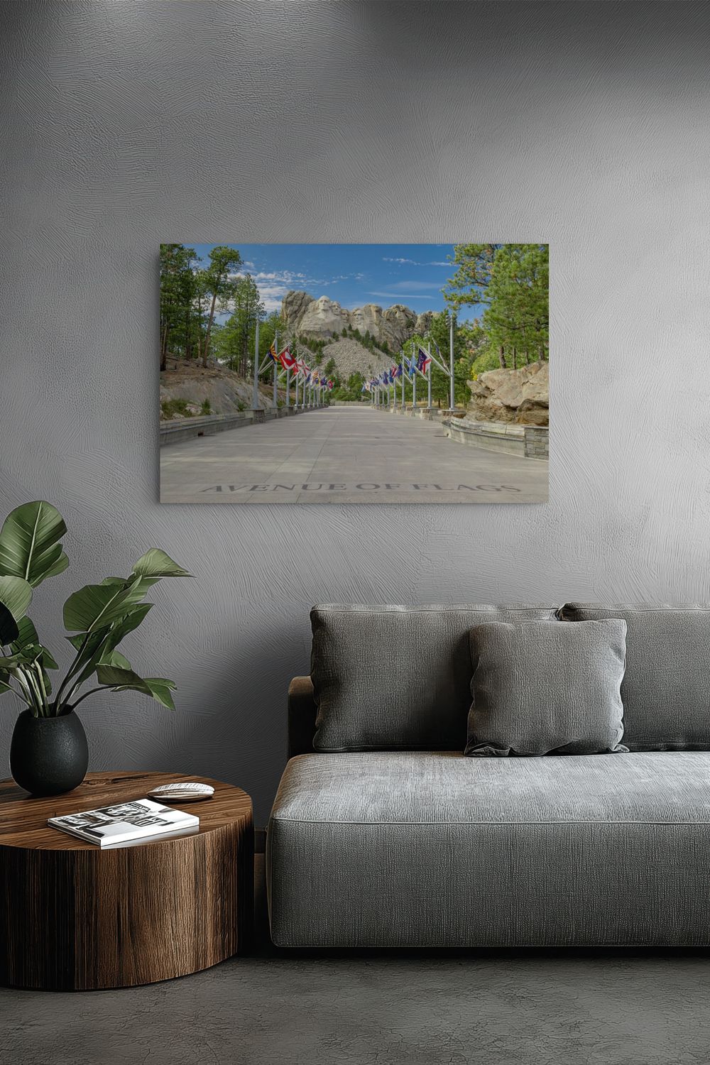 A rectangular wall art print showing a scenic road leading up a mountain with trees, flags, and rocky peaks under a blue sky, displayed above a gray sofa in a modern living room setting.