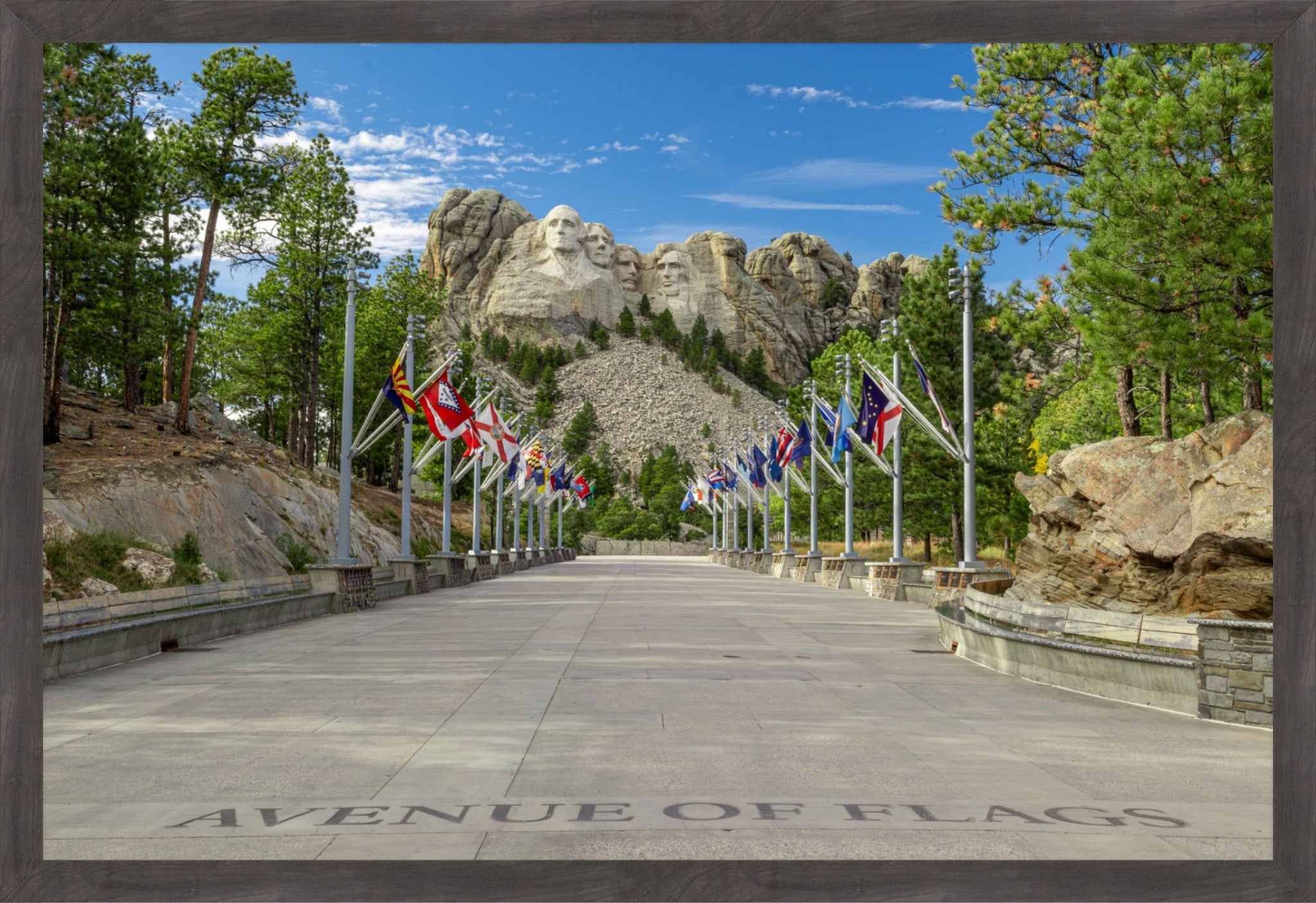 A framed scenic photograph of Mount Rushmore National Park, showing the Avenue of Flags pathway lined with flags, leading toward the rock formations under a blue sky with trees on both sides.