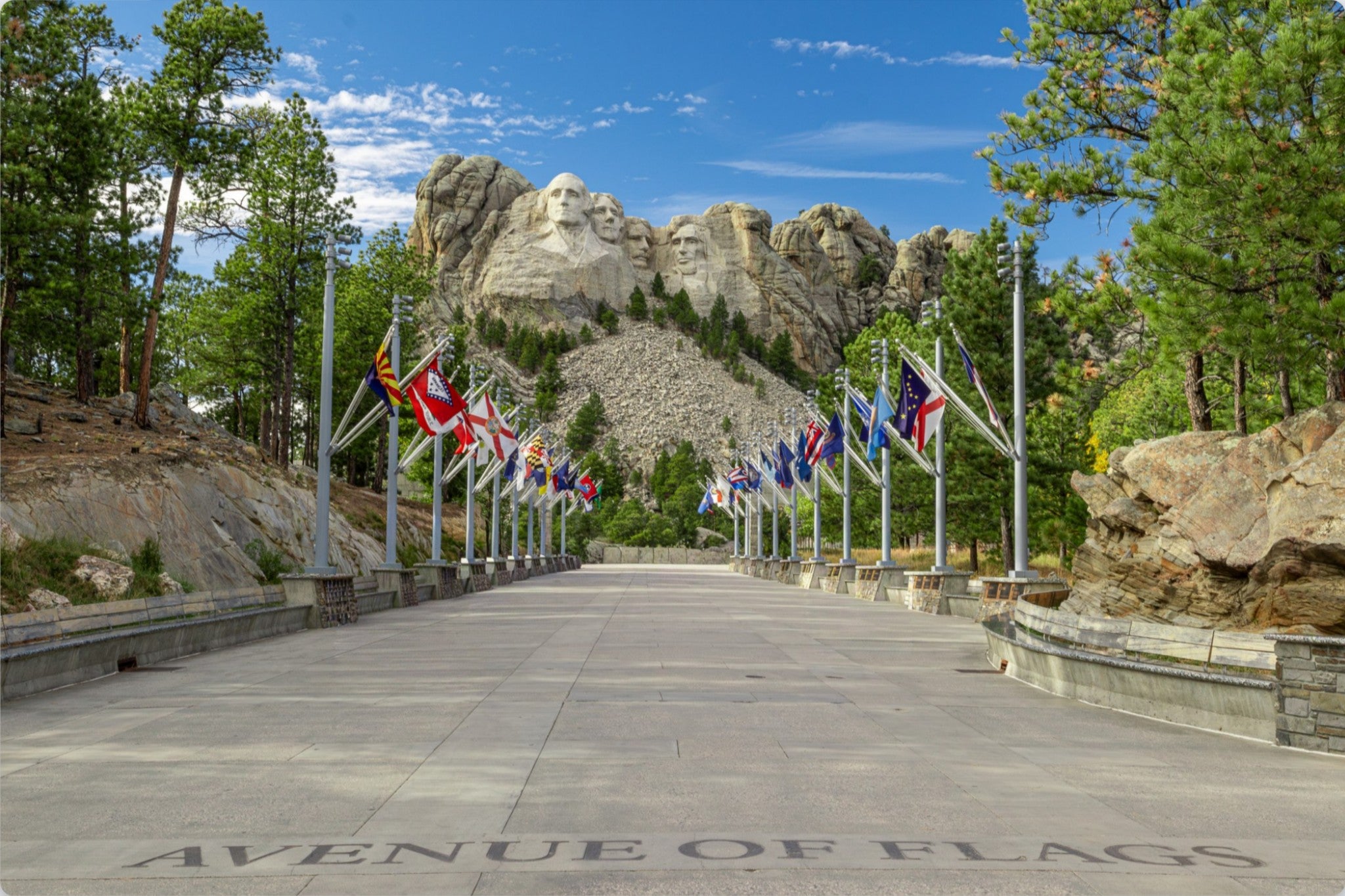 A paved pathway leading toward Mount Rushmore under a blue sky, lined with multiple international flags, trees, and rock formations. The words 'AVENUE OF FLAGS' are visible on the pavement.