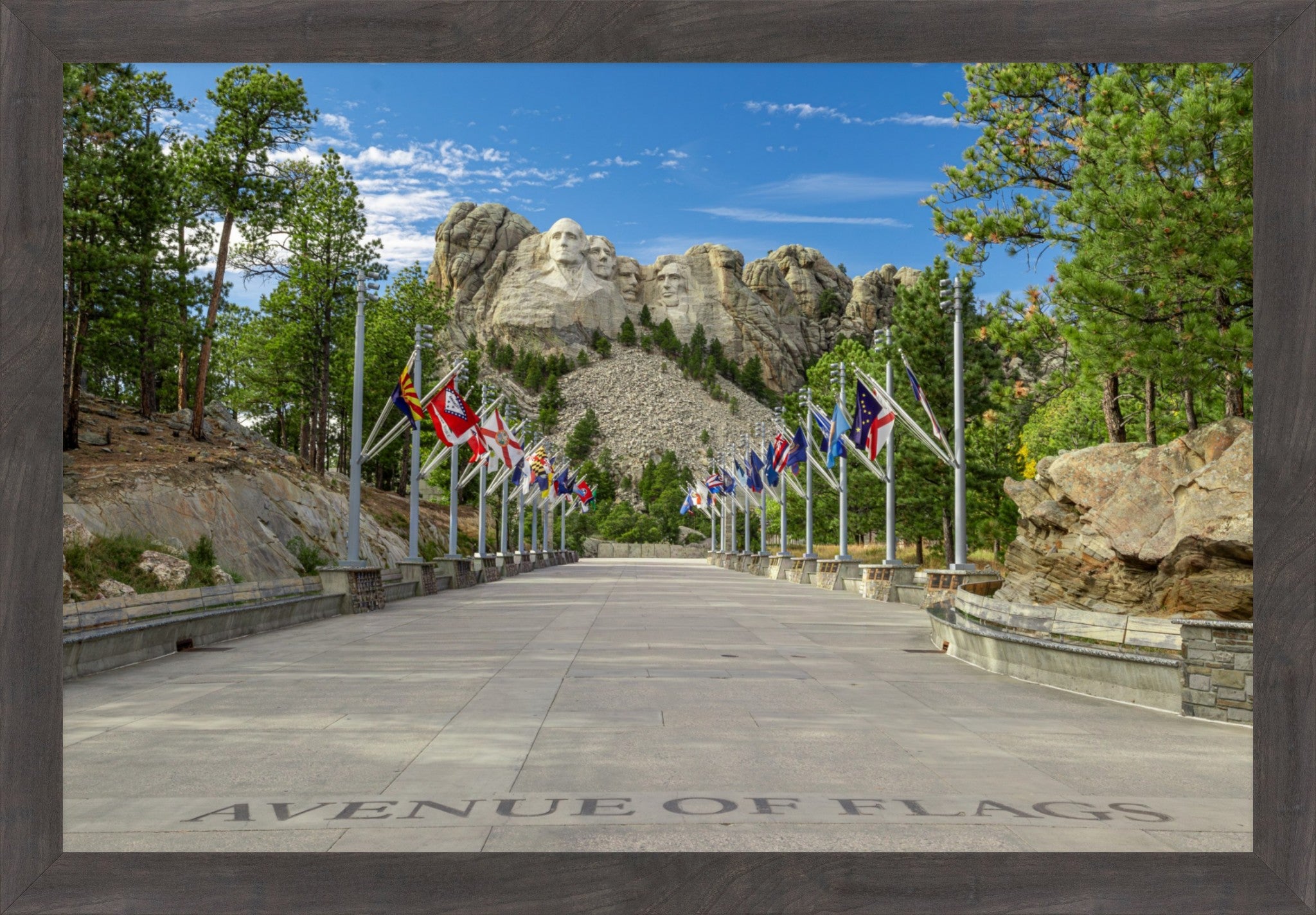 A framed photographic print of the Avenue of Flags leading up Mount Rushmore, with flags lining the path, trees, and rocky mountain peaks under a blue sky.