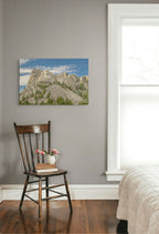 A landscape art print of a rocky mountain peak under a blue sky, displayed on a gray wall in a bedroom setting above a wooden chair and next to a window.