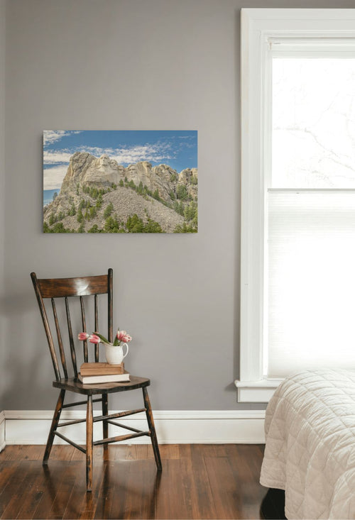 A landscape art print of a rocky mountain peak under a blue sky, displayed on a gray wall in a bedroom setting above a wooden chair and next to a window.