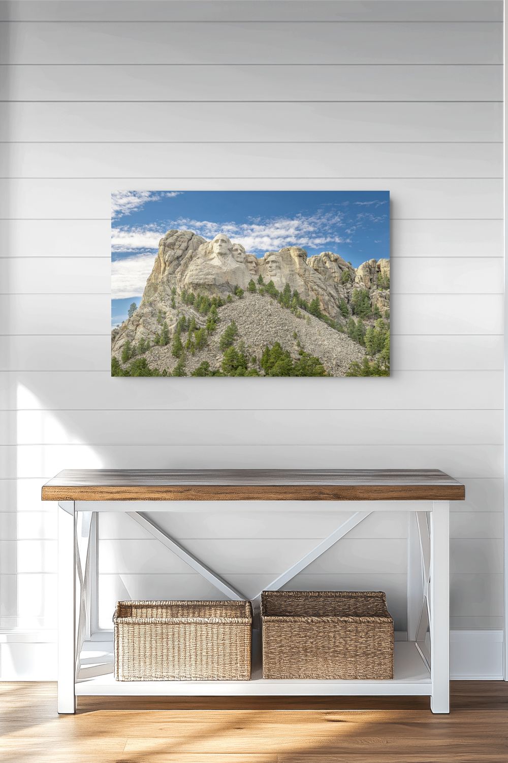 A rectangular wall art print showing a mountain landscape under a blue sky, displayed above a white console table with a wood top in a bright room with white shiplap walls and wood flooring.