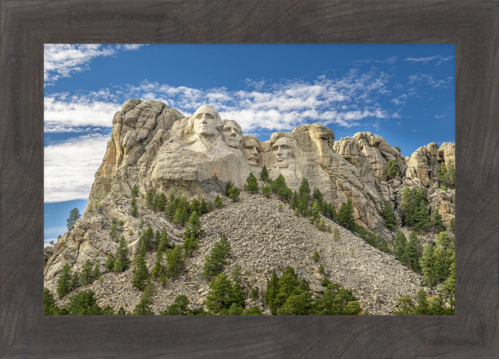 A framed landscape photograph of Mount Rushmore under a blue sky with white clouds, set in a dark gray wood frame.