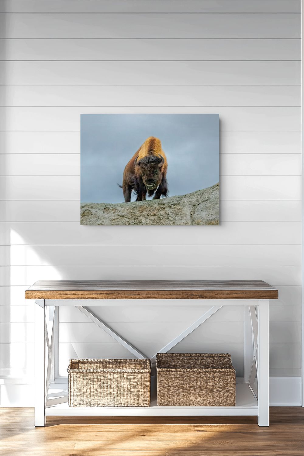 A square wall art print showing a bison standing on a rocky outcrop under a cloudy sky, displayed above a wooden bench with woven baskets against a white shiplap wall.