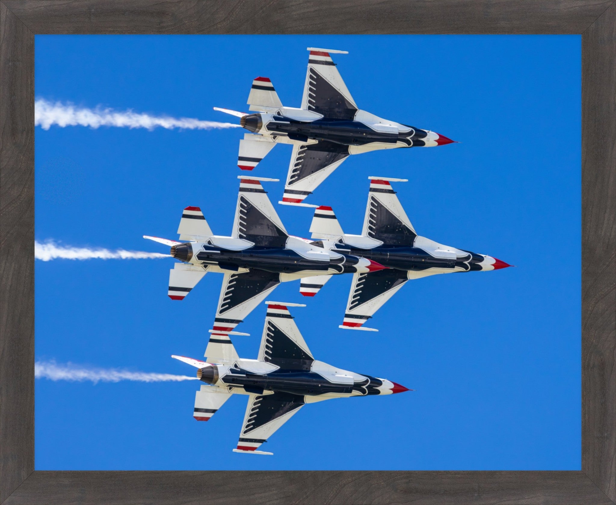 Framed wall art featuring four military jets flying in formation against a bright blue sky, with white smoke trails and red, white, and blue tail accents. The print is shown within a dark wood frame.