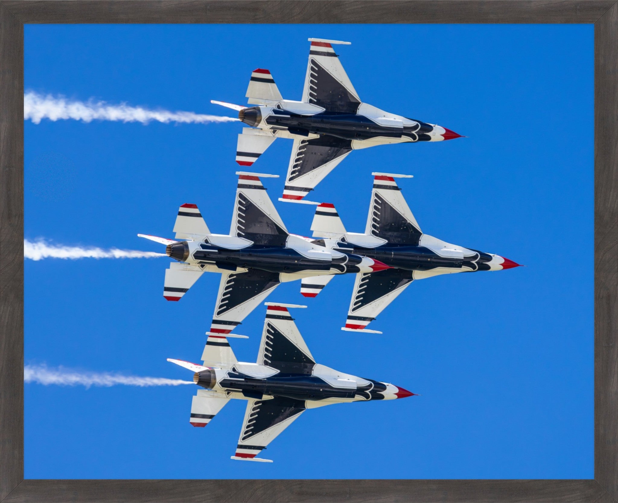 Framed wall art featuring a photograph of four military fighter jets flying in formation against a bright blue sky, with white contrails and red, white, and blue tail markings. The print is shown within a dark brown wood frame.
