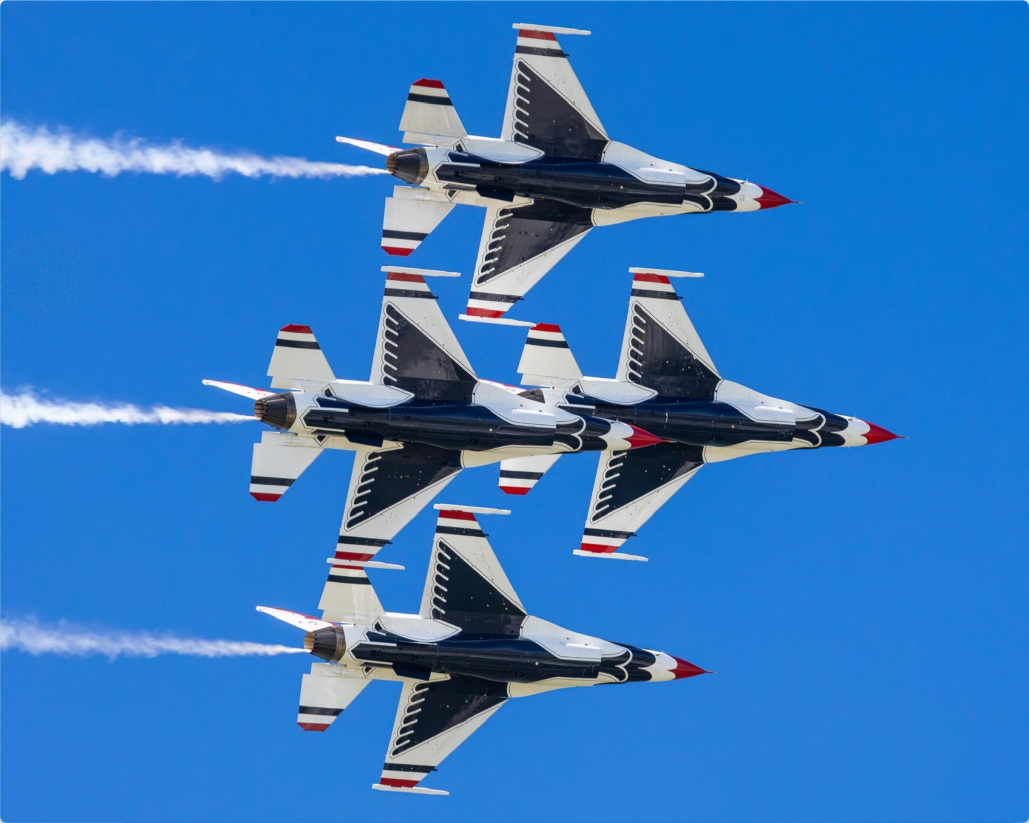 Four military jets flying in a tight formation against a clear blue sky, leaving white smoke trails.
