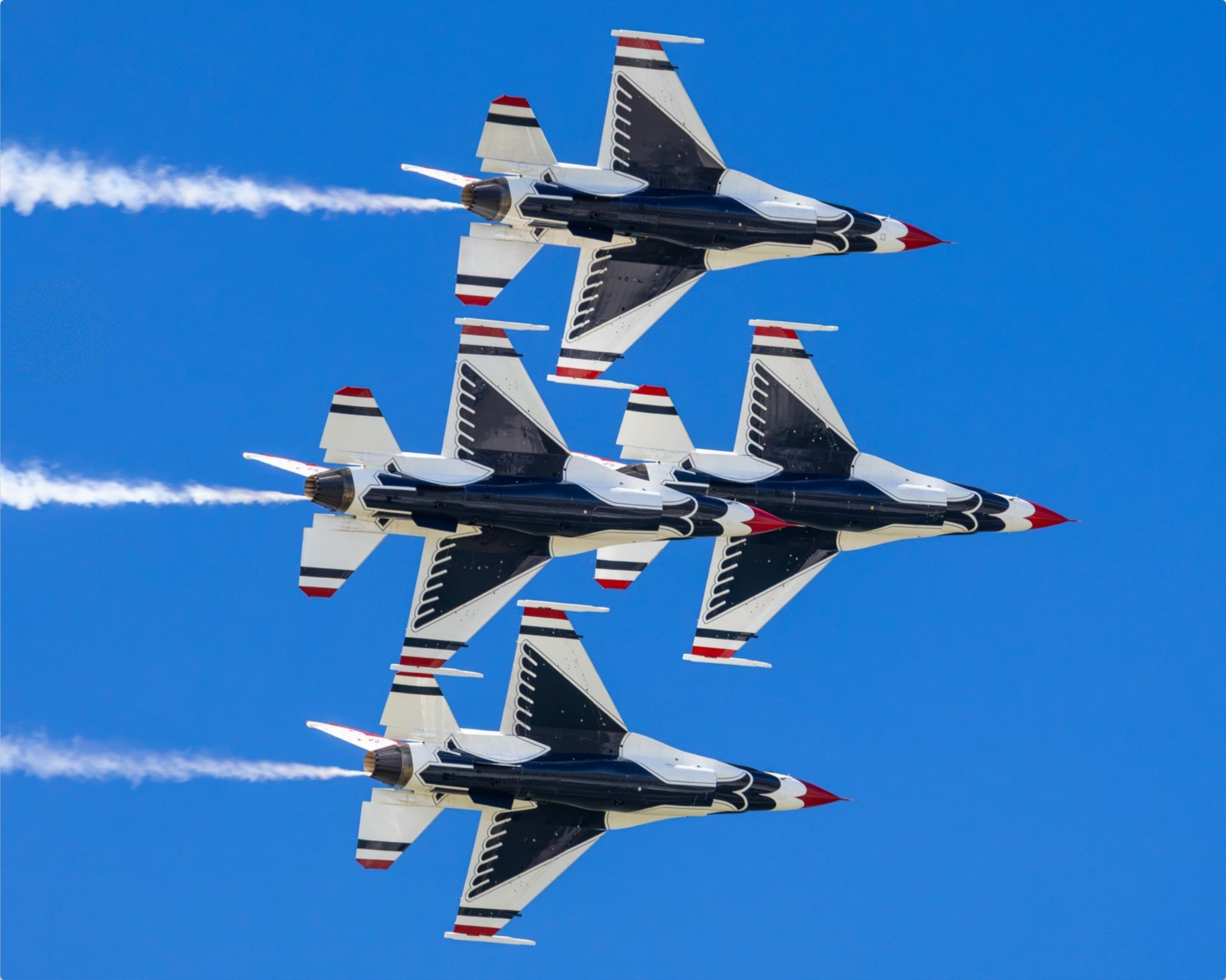 Four military jets flying in a tight formation against a clear blue sky, leaving white smoke trails.