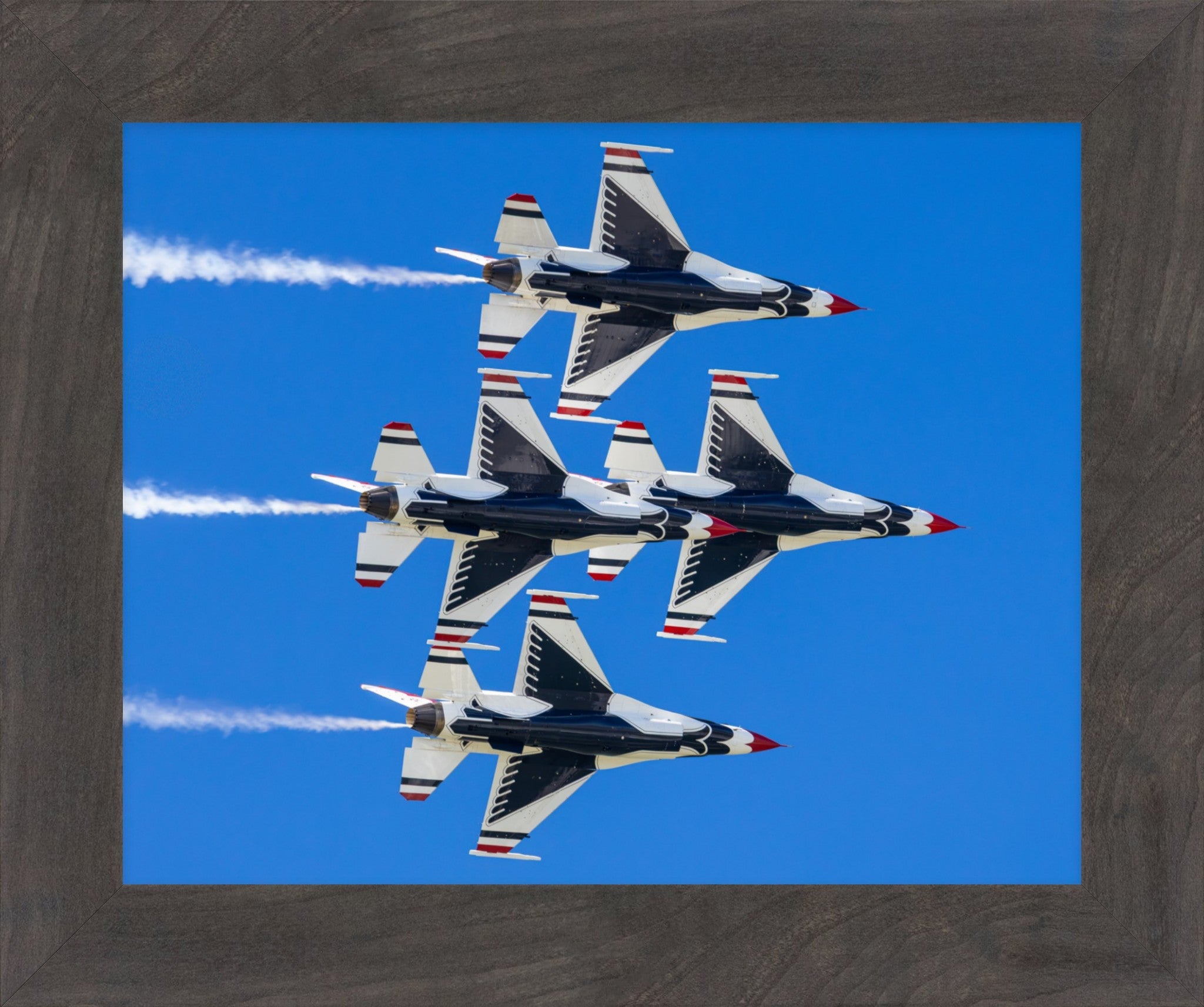 Framed wall art featuring four military jets flying in a V-formation against a bright blue sky, with white smoke trails and red, white, and blue tail accents. The print is housed in a dark brown wood-grain frame.