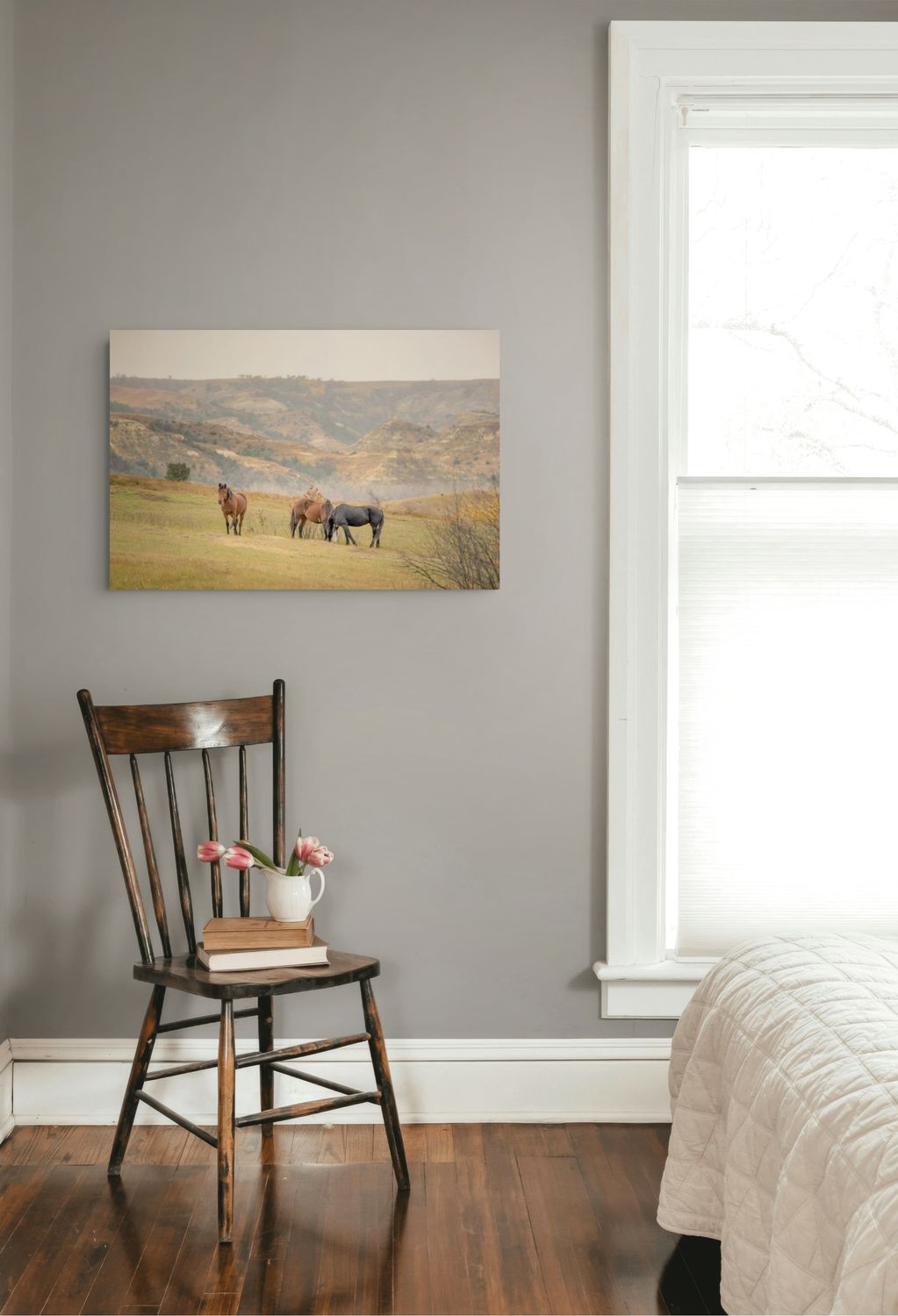A landscape art print depicting horses in a golden field under a hazy sky, displayed on a gray wall in a bedroom setting above a wooden chair and next to a window.