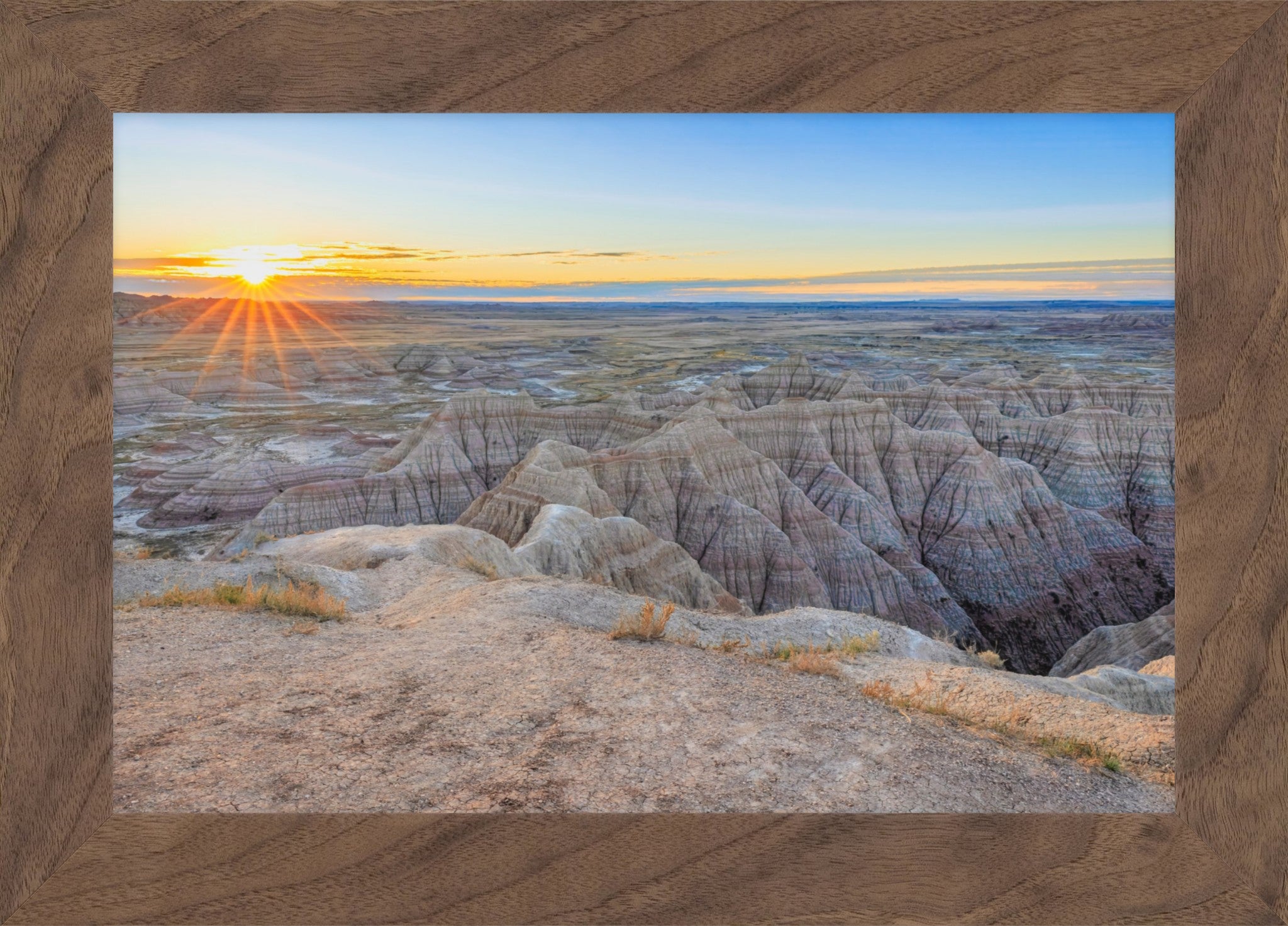 A framed landscape photograph of a badlands canyon at sunset, with layered rock formations in the foreground and a bright sun on the horizon under a blue sky, set within a rustic brown wood frame.