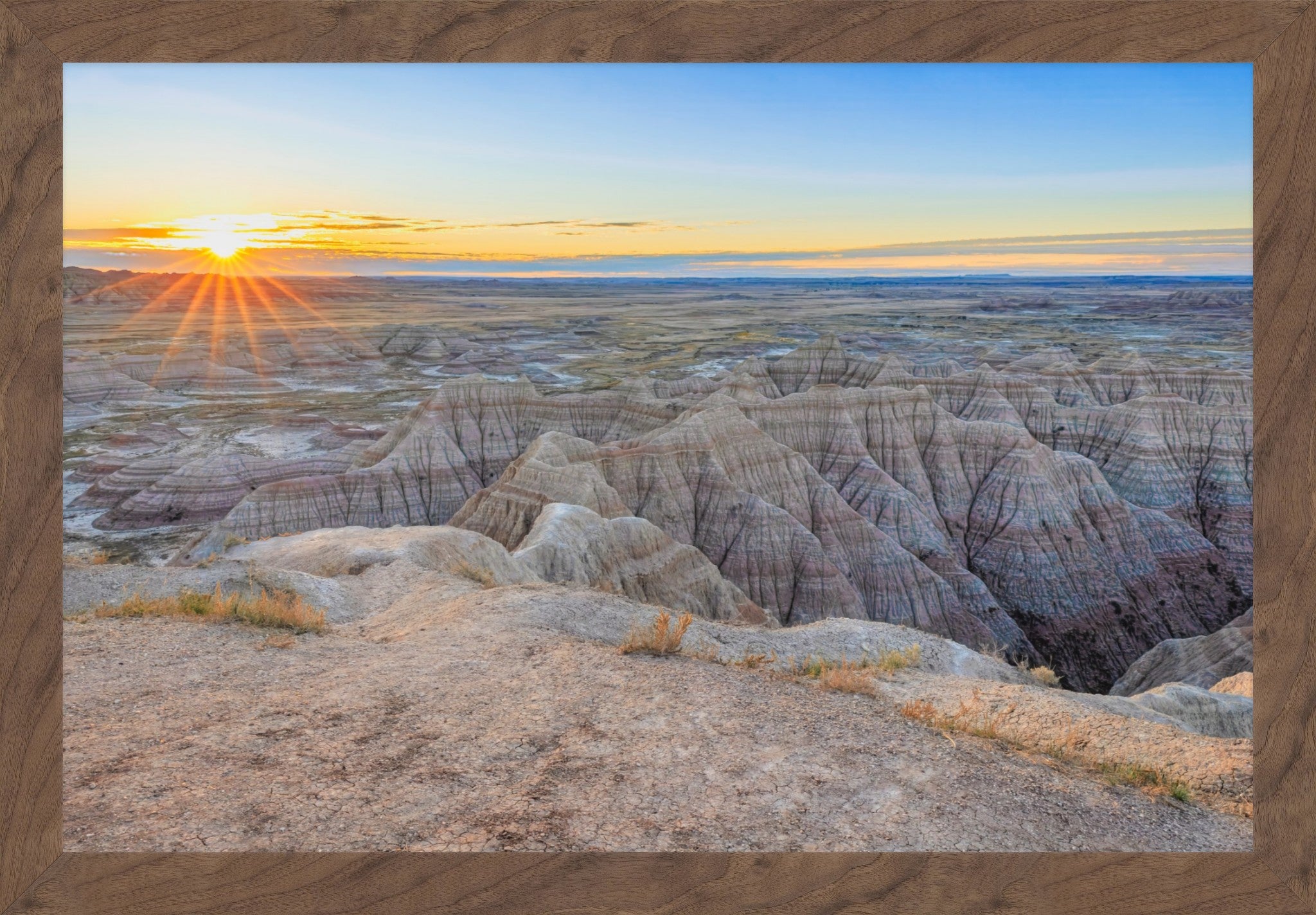 A framed landscape photograph of a badlands canyon at sunset, with layered rock formations in the foreground, a bright sunburst on the left, and a blue sky above, set within a brown wood frame.