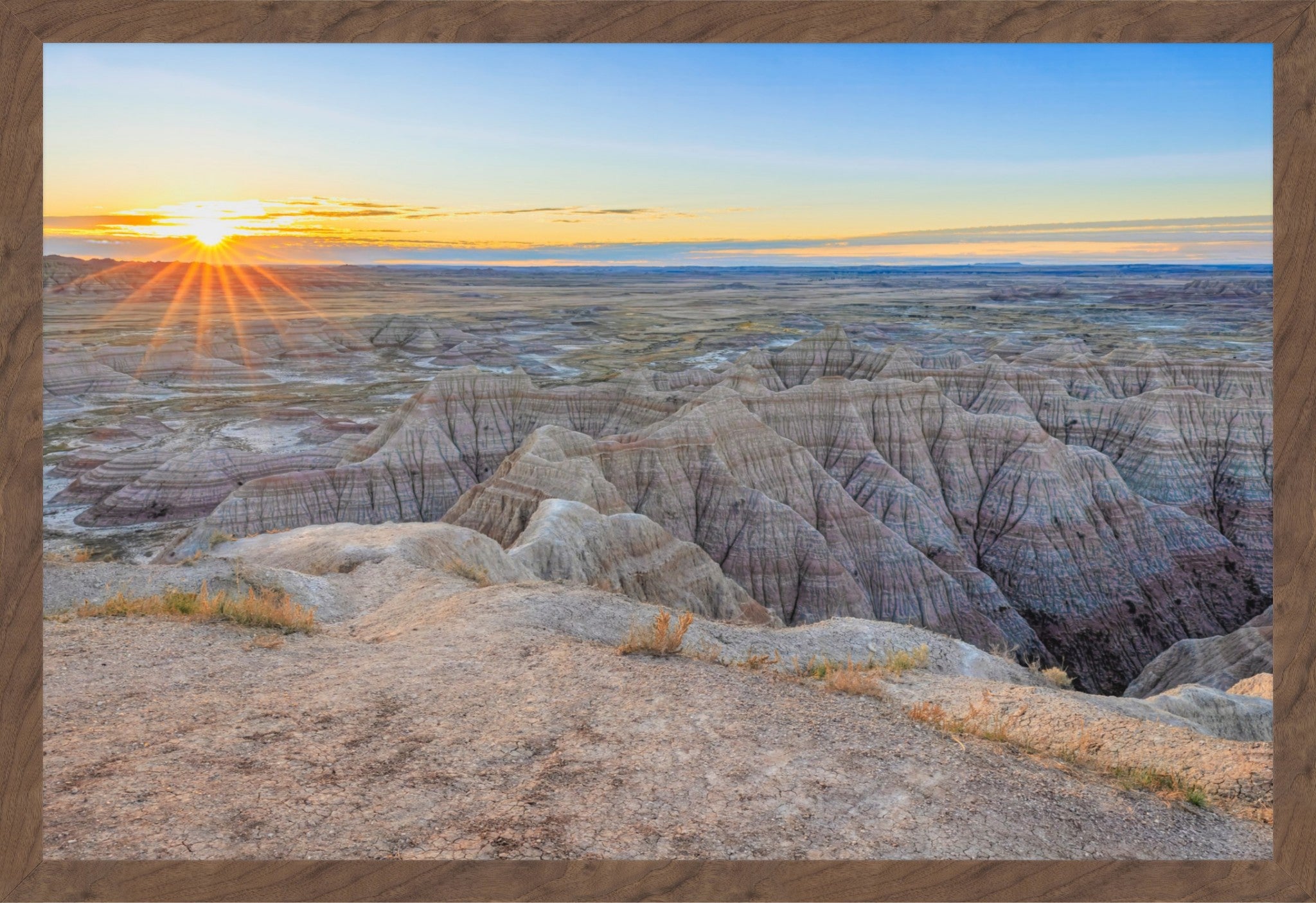 A framed landscape photograph of a sun setting over a badlands canyon with layered rock formations under a blue sky, bordered by a rustic wood frame.