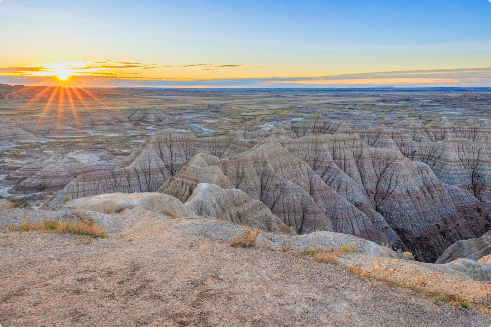 A scenic landscape photograph of a badlands canyon at sunset, showing layered beige and gray rock formations under a blue sky with the sun low on the horizon.