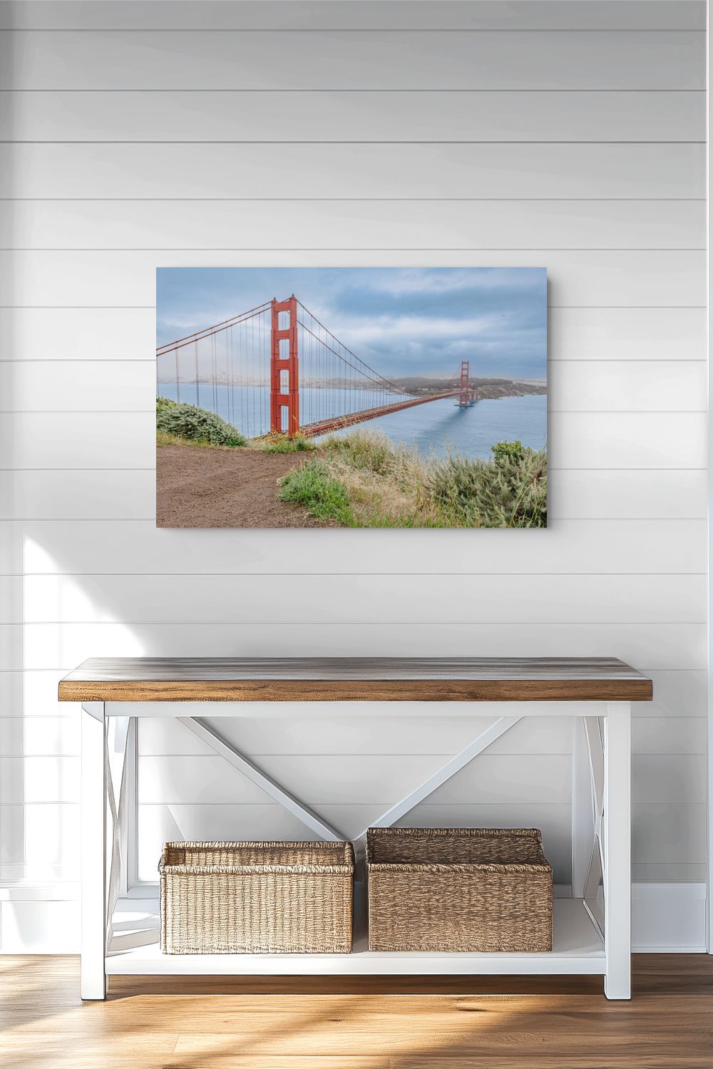A rectangular wall art print showing the Golden Gate Bridge over water, displayed above a white bench with a wood top in a bright room with white shiplap walls and wood flooring.