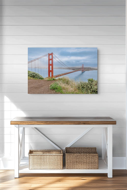 A rectangular wall art print showing the Golden Gate Bridge over water, displayed above a white bench with a wood top in a bright room with white shiplap walls and wood flooring.