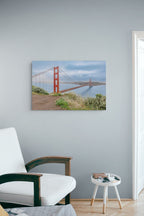 A rectangular wall art print showing the Golden Gate Bridge over water, displayed on a light gray wall above a white chair and a small side table in a styled interior.