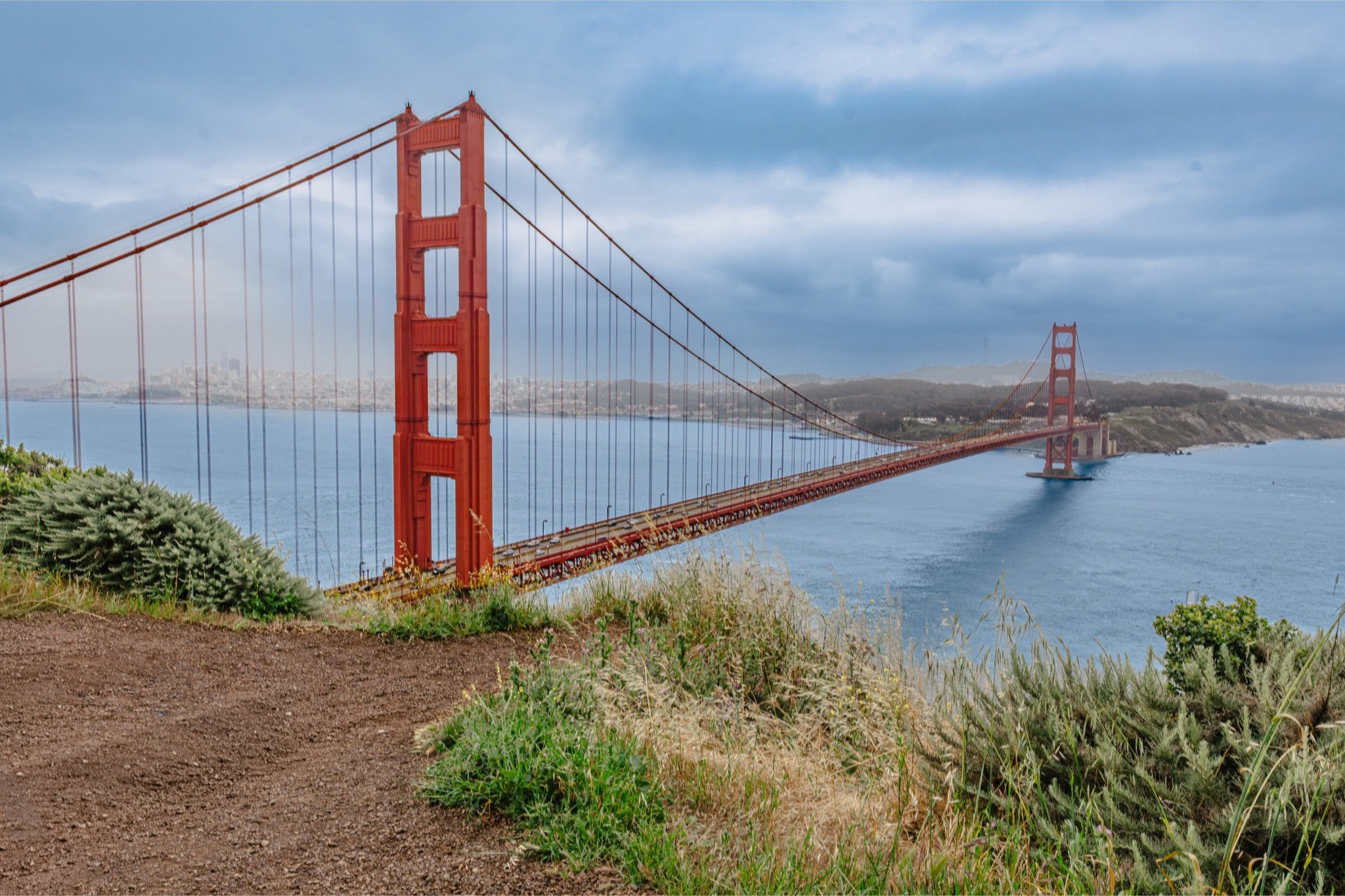 The Golden Gate Bridge spans across blue water under a cloudy sky, with green and brown vegetation in the foreground.