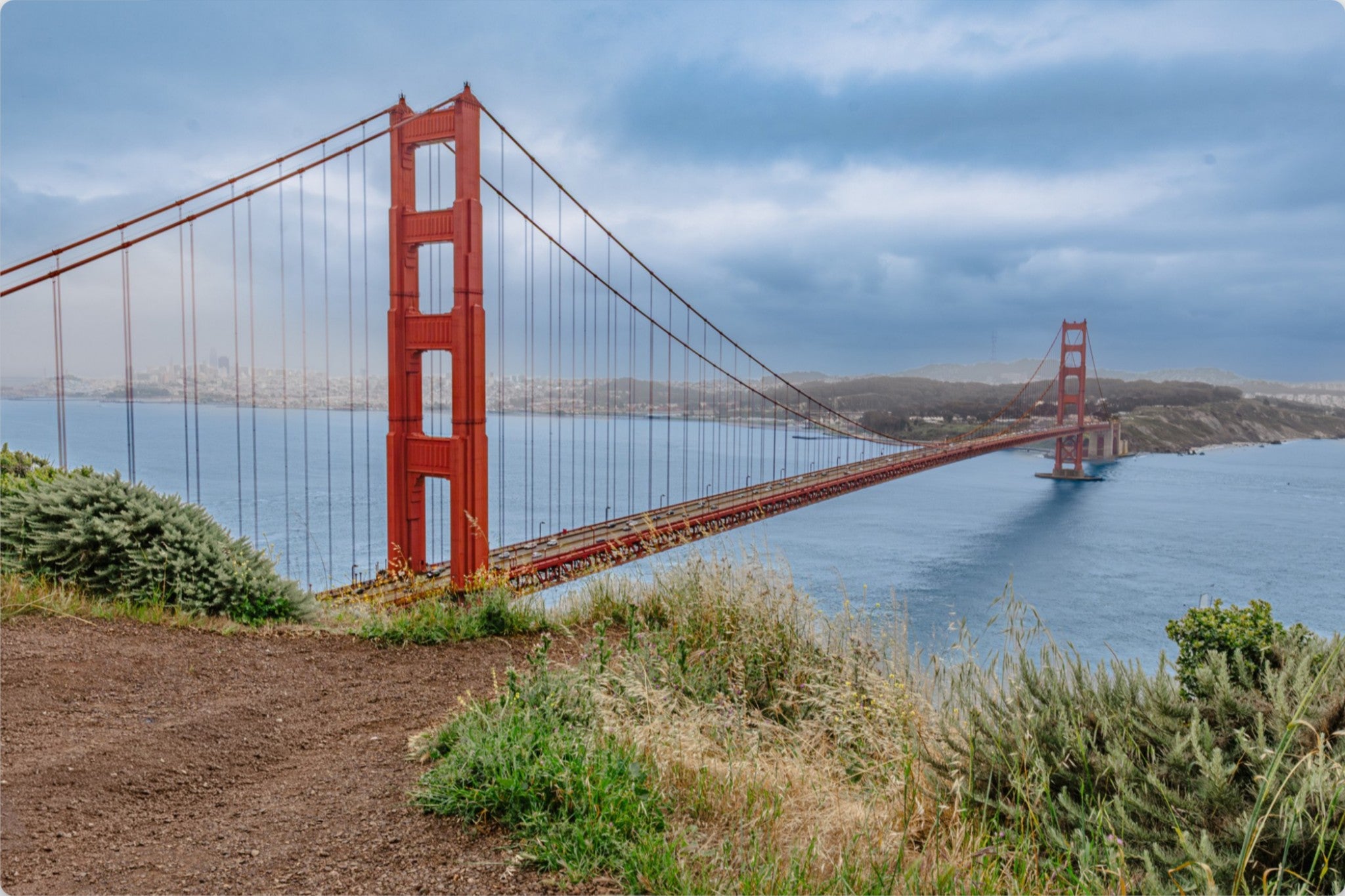 The Golden Gate Bridge spans the bay under a cloudy blue sky, with blue water, distant hills, and foreground vegetation visible.