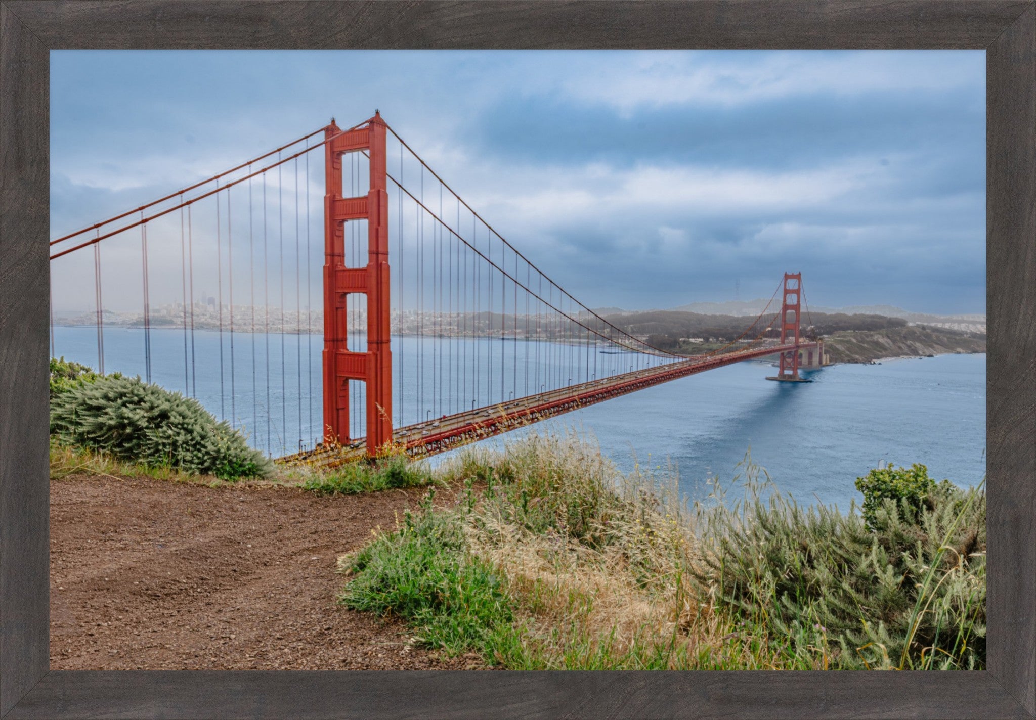 A framed photographic print of the Golden Gate Bridge over the water, with dramatic clouds in the sky and grassy foreground, surrounded by a dark wood frame.