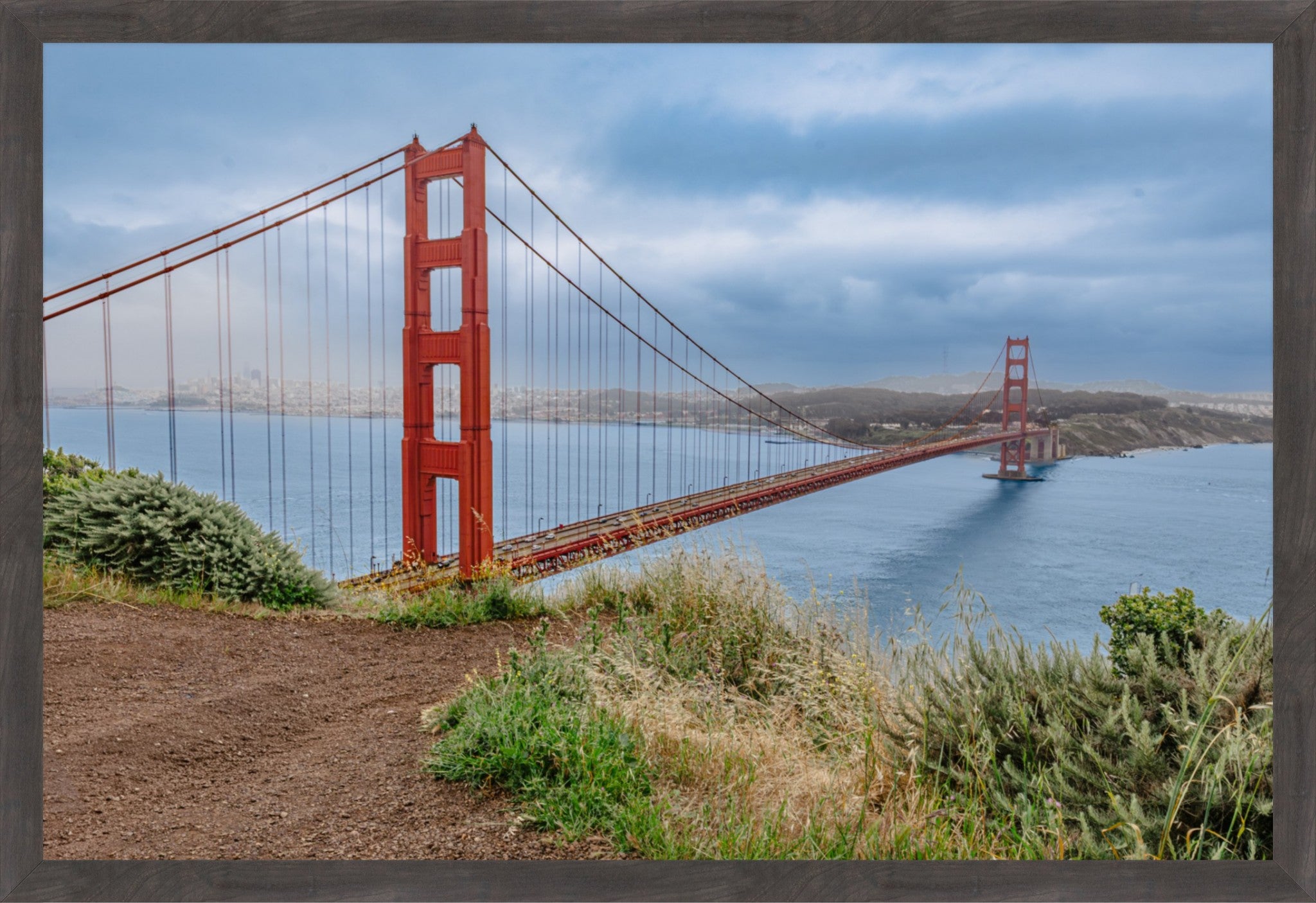 A framed photographic print of the Golden Gate Bridge over blue water under a cloudy sky, with grassy foreground and a dark gray frame border.