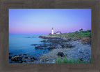 A framed coastal landscape print showing a lighthouse on a rocky shoreline at dusk, with blue water, purple and pink sky, and a dark wood frame around the artwork.