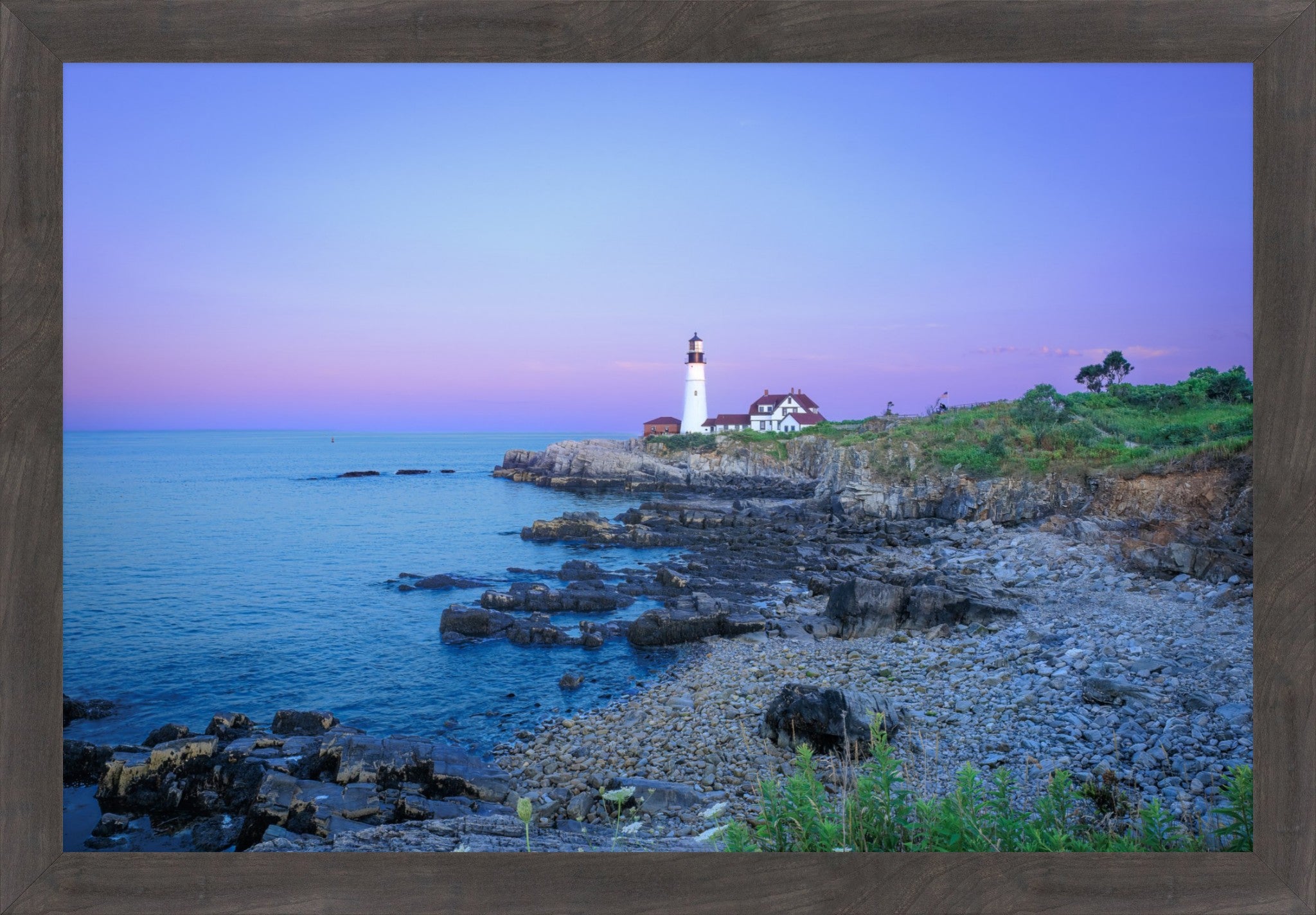 A framed coastal landscape print showing a lighthouse on a rocky shoreline at dusk, with blue water, purple sky, and green vegetation, surrounded by a brown wood frame.