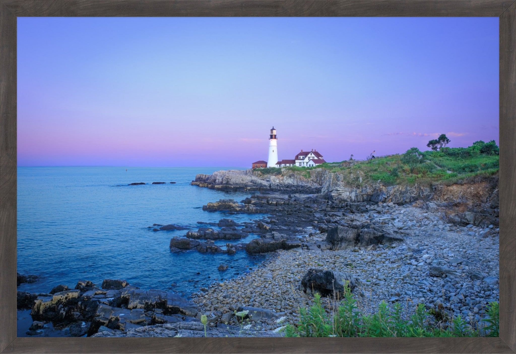 A framed coastal landscape print showing a lighthouse on a rocky shoreline beside calm blue water under a purple and pink sky, enclosed in a dark wood frame.
