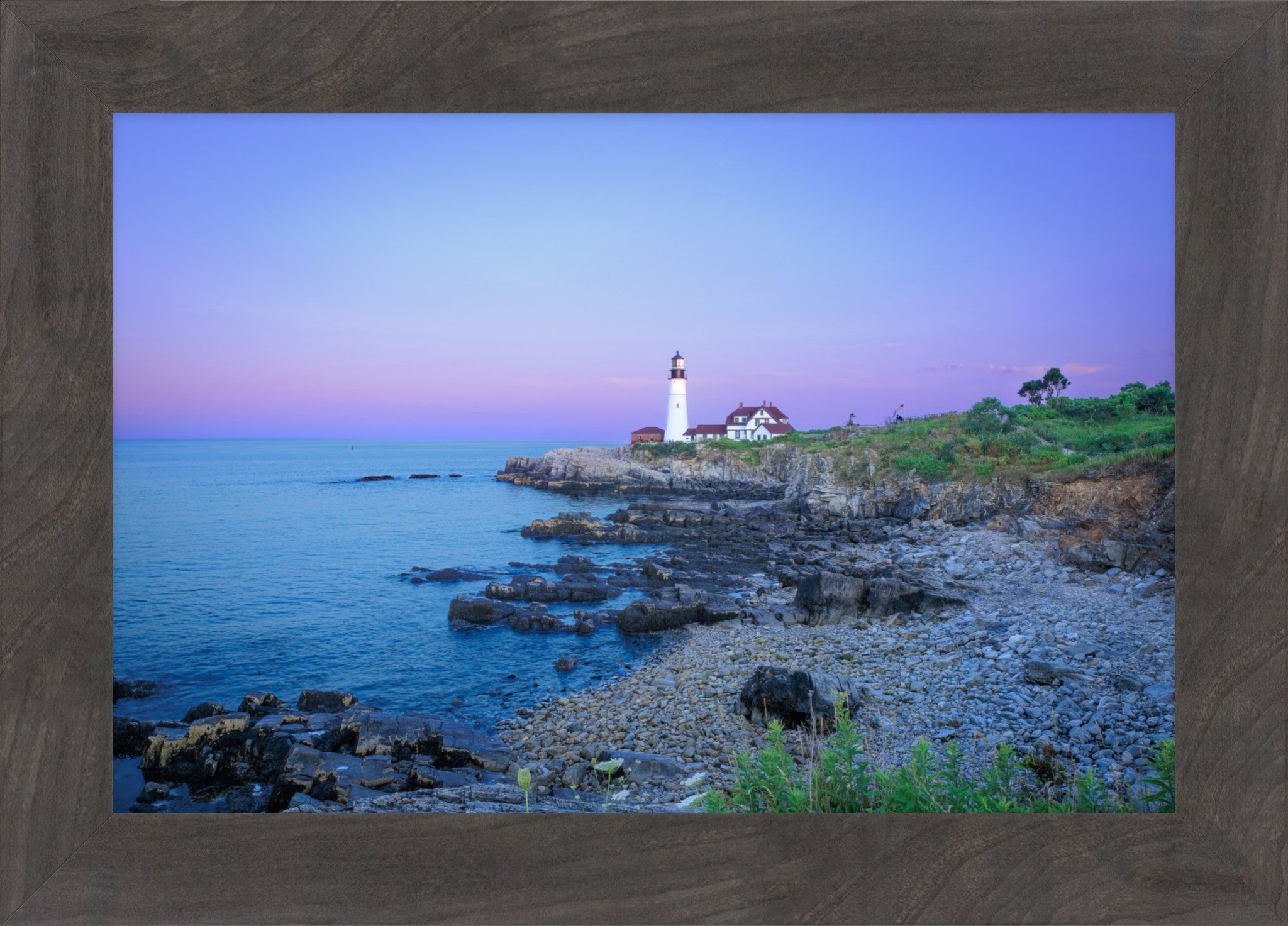 A framed coastal landscape print showing a lighthouse on a rocky shoreline at dusk, with blue water, purple and pink sky, and a brown wood-look frame.
